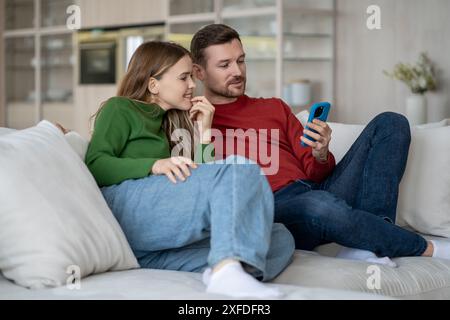 Joyful married couple smiling, laughing sitting on couch with phone browsing online internet content Stock Photo