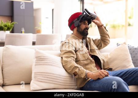 Using VR headset, Indian man sitting on couch in modern living room, copy space Stock Photo