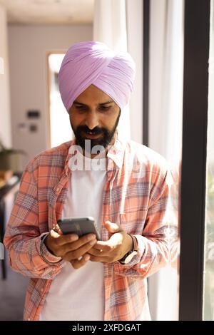 Using smartphone, Indian man in turban standing by window at home Stock Photo