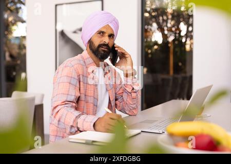 Talking on smartphone, Indian man writing in notepad and using laptop at home Stock Photo