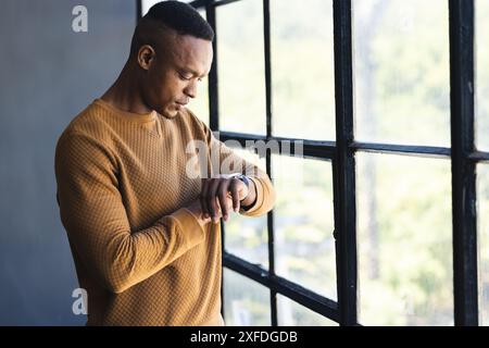Checking smartwatch, man standing by window in casual sweater, focusing on time Stock Photo