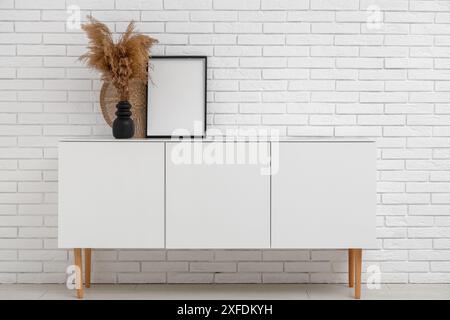 Cabinet with empty frame and pampas grass in vase near white brick wall Stock Photo