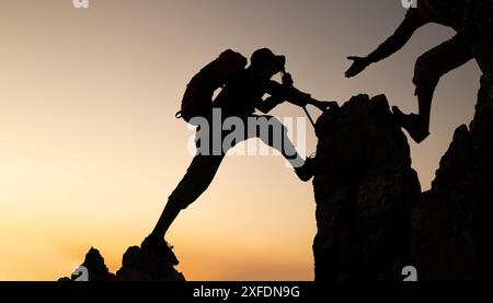 silhouette of two male climbers scaled the cliff and one of them offered a helping hand. person who help and work as a team. Stock Photo