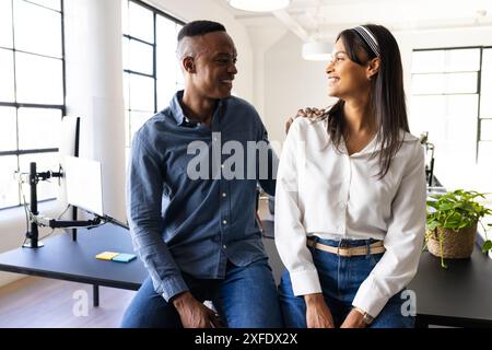 Smiling colleagues sitting on desk in modern office, enjoying conversation Stock Photo