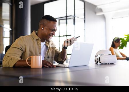 Smiling man using smartphone and laptop in modern office with coffee mug Stock Photo