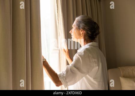 Looking out window, mature woman in white shirt enjoying sunlight at home, copy space Stock Photo