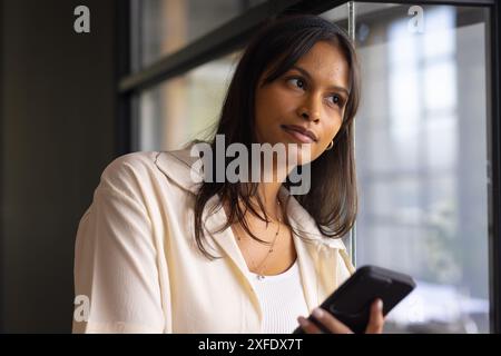 Holding smartphone, businesswoman leaning on window, thinking in modern office Stock Photo