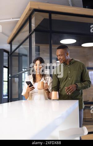 Discussing work, colleagues holding coffee mugs and using smartphone in office Stock Photo