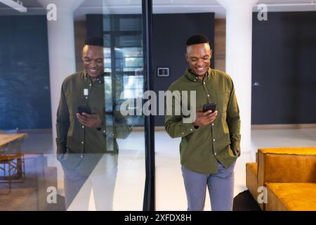 Smiling businessman using smartphone while standing by glass wall in modern office, copy space Stock Photo