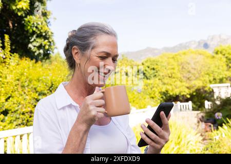 Smiling mature woman holding coffee mug and using smartphone outdoors Stock Photo