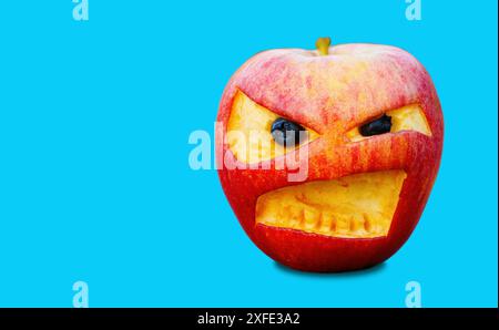 Red apple with carved angry-looking eyes and a frowning mouth isolated on blue. Stock Photo