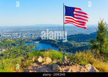 Patriotic scene featuring an American flag fluttering in the breeze above panoramic views of Los Angeles cityscape and lush hills from Mount Lee on a Stock Photo