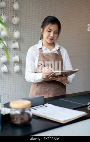 An attractive young Asian female barista or waitress is working in a coffee shop, writing orders on the paper. service, hospitality, small business Stock Photo
