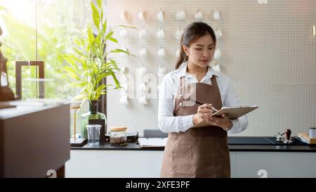 An attractive young Asian female barista or waitress is working in a coffee shop, writing orders on the paper. service, hospitality, small business Stock Photo