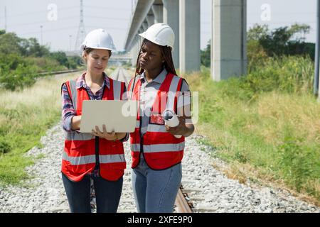 Two engineer with safety helmet and high-visibility vest reviewing work to be done on a railway track project while holding laptop computer and a set Stock Photo