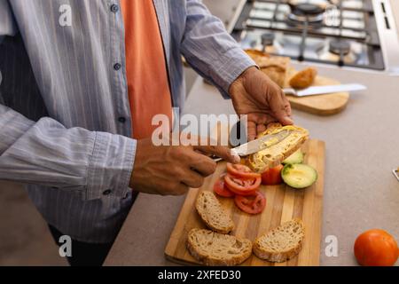 Spreading butter on bread, man preparing sandwich with fresh vegetables in kitchen Stock Photo