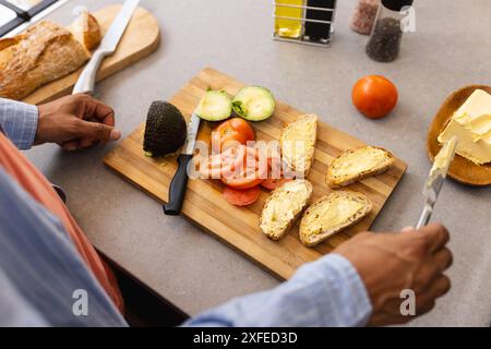 Preparing breakfast, man spreading butter on bread with avocado and tomato slices Stock Photo