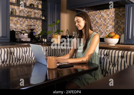 Working from home, young woman using laptop at kitchen table Stock Photo