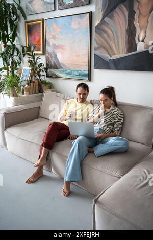 Pleased married couple sitting on sofa with laptop at art home studio, spending weekend together. Stock Photo
