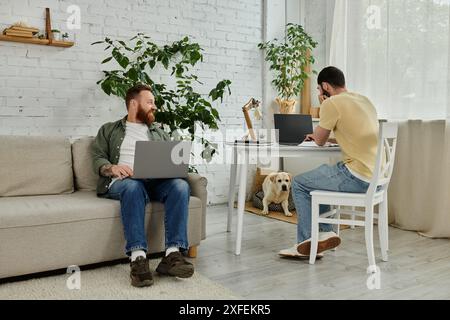 A couple of men are sitting on top of a couch in a living room, working remotely with a labrador dog nearby. Stock Photo