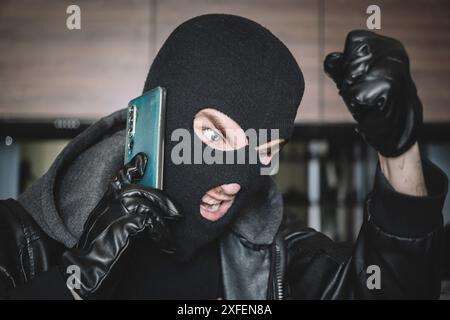 The thief is talking on phone in a black mask. extortionist demands money for the kidnapping. a phone fraudster commits a crime, violates the law. con Stock Photo