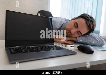 Overworked young freelancer man sitting at workplace and sleeping, napping while working on laptop computer, placed head on hand, feeling tired, copy Stock Photo