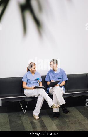Male and female medical colleagues discussing over documents while sitting on chair near wall in hospital Stock Photo