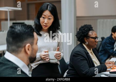 Female business professional holding coffee cup while discussing with male colleague in office Stock Photo