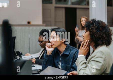 Thoughtful businessman leaning on elbow while looking at computer screen and sitting with female coworker in office Stock Photo