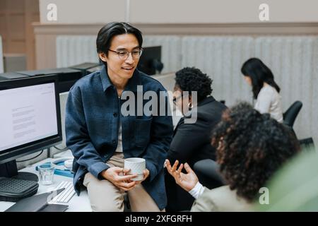 Businesswoman holding coffee cup while sitting on desk and discussing with female colleague at office Stock Photo