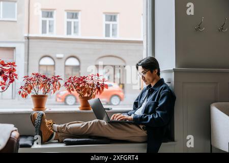 Businessman working on laptop while sitting on window sill at office Stock Photo