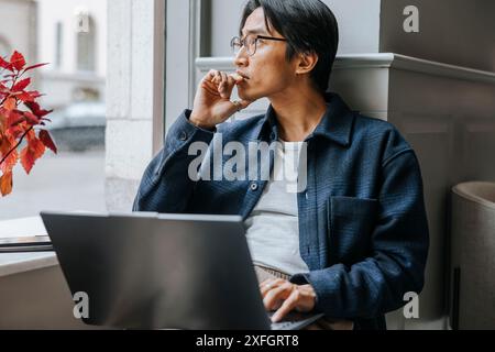 Thoughtful businessman sitting with laptop on window sill at office Stock Photo