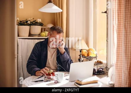Happy mature man enjoying watching laptop while sitting at dining table near window Stock Photo