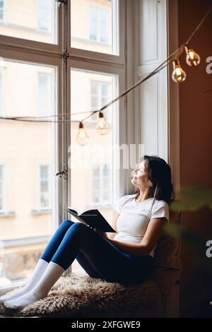 Young woman sitting with book on window sill at home Stock Photo