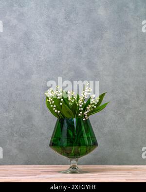 A small bouquet of white lilies of  valley in a green transparent vase in  form of a glass. copy space Stock Photo