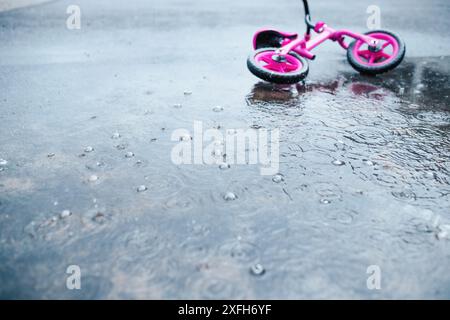A pink child's bicycle sits in a puddle of rainwater, with raindrops creating ripples on the surface. Stock Photo