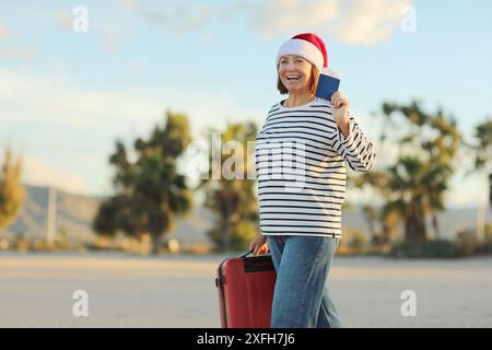 Excited cheerful adult woman in Christmas Santa Claus hat holds red suitcase, passport and boarding ticket walks on beach on seashore with palms, celebrates new year holidays vacation at exotic resort Stock Photo