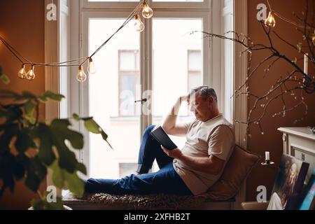 Mature man reading book while sitting on window sill at home Stock Photo