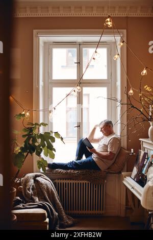 Mature man reading book sitting near window at home Stock Photo