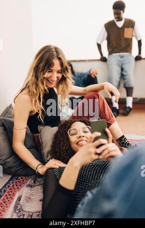 Smiling woman sitting near female friend using smart phone on building terrace Stock Photo