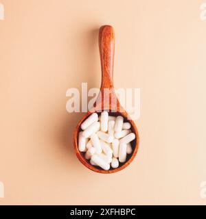 Top view of wooden spoon filled with white capsules on beige background, square composition. Minimalist healthcare and wellness concept. Stock Photo