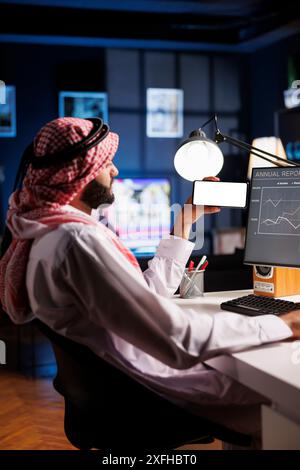 Side-view shot Middle Eastern man in traditional attire sitting at an office desk, using a desktop computer. They hold a mobile phone with a blank chromakey mockup template, offering space for advertising. Stock Photo