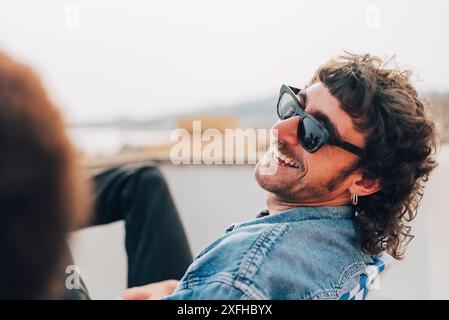 Happy man wearing sunglasses and laughing while sitting on building terrace Stock Photo