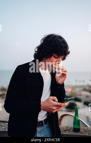 Young man eating pizza and using smart phone standing at rooftop Stock Photo
