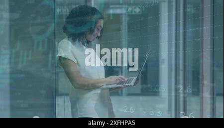 Image of graphs, trading board, loading circles, biracial woman standing and using laptop Stock Photo