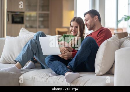 Happy married couple, husband, wife sitting relaxed on couch using laptop, laughing, smiling. Stock Photo