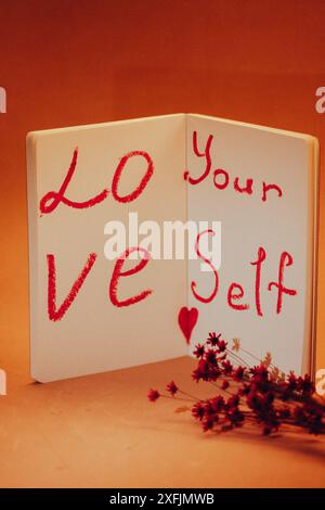 Text 'Love yourself' written in red lipstick on white sheets of open notebook Self-love, healthy narcissism concept A bouquet of small dry red flowers Stock Photo