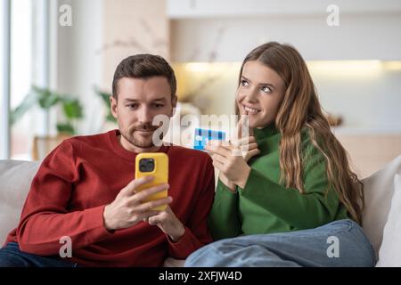 Happy married couple spouses shopping online via internet using smartphone, sitting on couch. Stock Photo