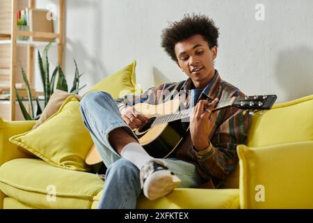 A young African American man playing an acoustic guitar on a yellow couch while talking on his phone. Stock Photo