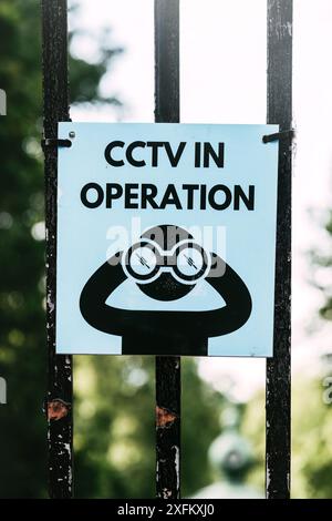 A blue and black sign with the words CCTV In Operation and a cartoon person with binoculars, attached to a black metal gate in London Stock Photo